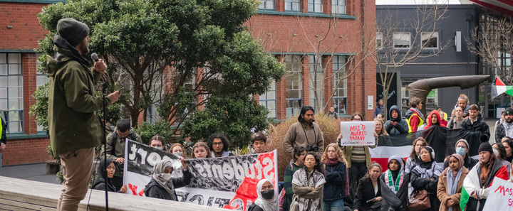 Will is standing on a platform holding a microphone addressing a Palestine rally Will is standing on a platform holding a microphone addressing a Palestine rally